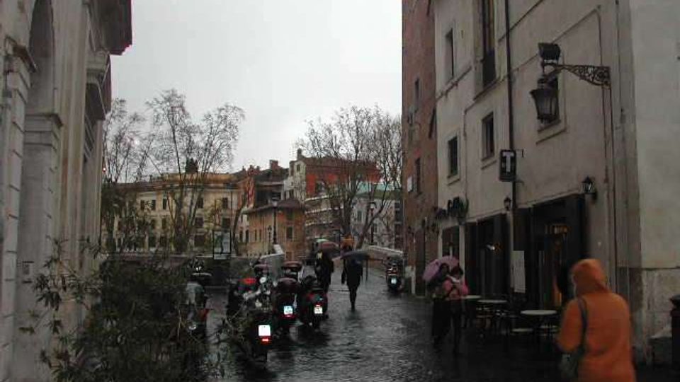 Strada urbana in pavé bagnato in un centro storico, fiancheggiata da edifici alti con facciate chiare e lampioni a parete. Sul lato destro si vedono tavolini all’aperto e una fila di scooter parcheggiati, con alberi spogli e palazzi sullo sfondo.
