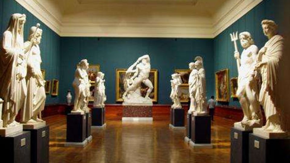 Sala di museo con pareti verde scuro e lucernario centrale, pavimento in legno lucido e sculture in marmo su piedistalli disposte lungo il percorso. Dipinti incorniciati alle pareti e una grande scultura al centro della sala.