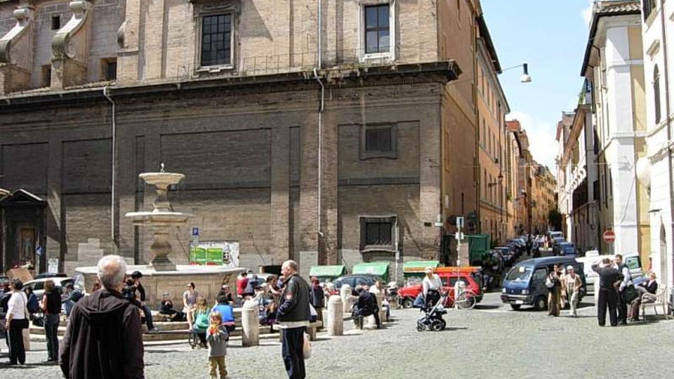 Piazza lastricata in sampietrini con una fontana in pietra al centro, circondata da edifici storici in muratura con grandi facciate e finestre alte. Sullo sfondo una strada stretta fiancheggiata da palazzi e auto parcheggiate, con lampioni e cartelli stradali.