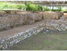 Scavi archeologici all’aperto con muri in pietra a bassa altezza e percorsi tra i resti, coperti da una tettoia metallica sostenuta da pilastri. Vegetazione e alberi circondano l’area sullo sfondo.