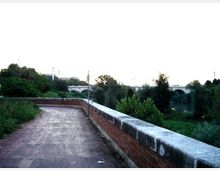 Percorso pedonale in cemento con muretto in mattoni e parapetto in pietra, affiancato da vegetazione. Sullo sfondo si vede un ponte ad archi sopra un fiume, circondato da alberi.