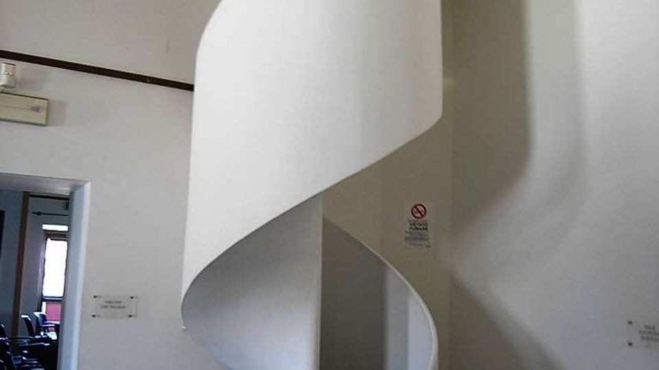 Scala a chiocciola in metallo verniciato bianco che sale verso un’apertura nel soffitto, collocata in un piccolo atrio interno con pareti chiare e pavimento in pietra. Accanto alla scala si vede un grande vaso in terracotta e una porta che conduce a un’altra stanza.