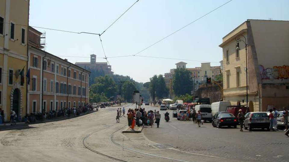 Ampia piazza urbana con binari del tram che curvano in primo piano e cavi elettrici sospesi sopra la strada. Ai lati si vedono edifici a più piani con facciate colorate e finestre, marciapiedi con veicoli parcheggiati e, sullo sfondo, una collina con un edificio storico.