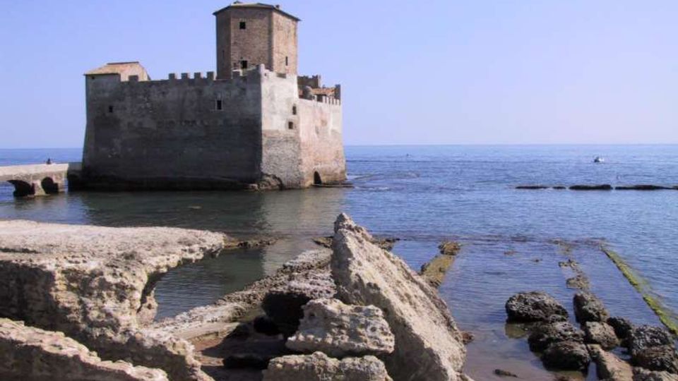 Castello in pietra affacciato sul mare, con torre centrale e mura merlate, costruito su un piccolo isolotto vicino alla riva. In primo piano si vedono scogli e rovine in pietra lungo la costa e acque calme sotto un cielo sereno.