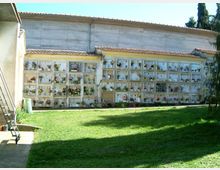 Cortile di un cimitero con prato verde e un lungo muro di loculi in cemento, disposti in file regolari e decorati con vasi di fiori. Sullo sfondo si vede l’edificio con tetto in tegole e alcuni alberi ai lati.
