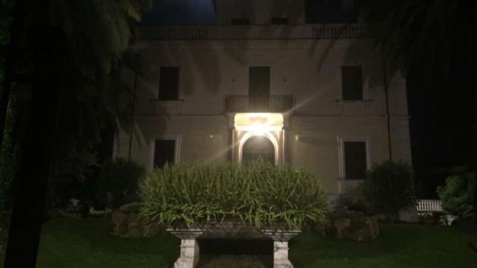 Facciata di una villa a due piani vista di notte, con ingresso ad arco illuminato al centro e finestre simmetriche. Davanti si trova un giardino con prato, un’aiuola rialzata e palme ai lati.