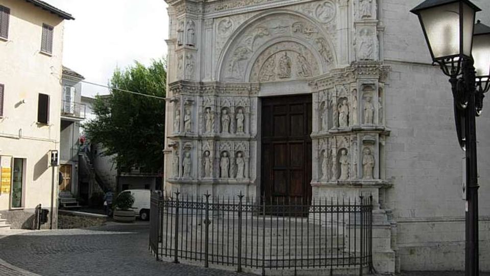 Facciata in pietra di una chiesa storica con portale ad arco riccamente scolpito e nicchie con statue, chiusa da una cancellata in ferro davanti a una scalinata. La chiesa si affaccia su una piccola piazza lastricata, con edifici bassi ai lati e un lampione in primo piano.