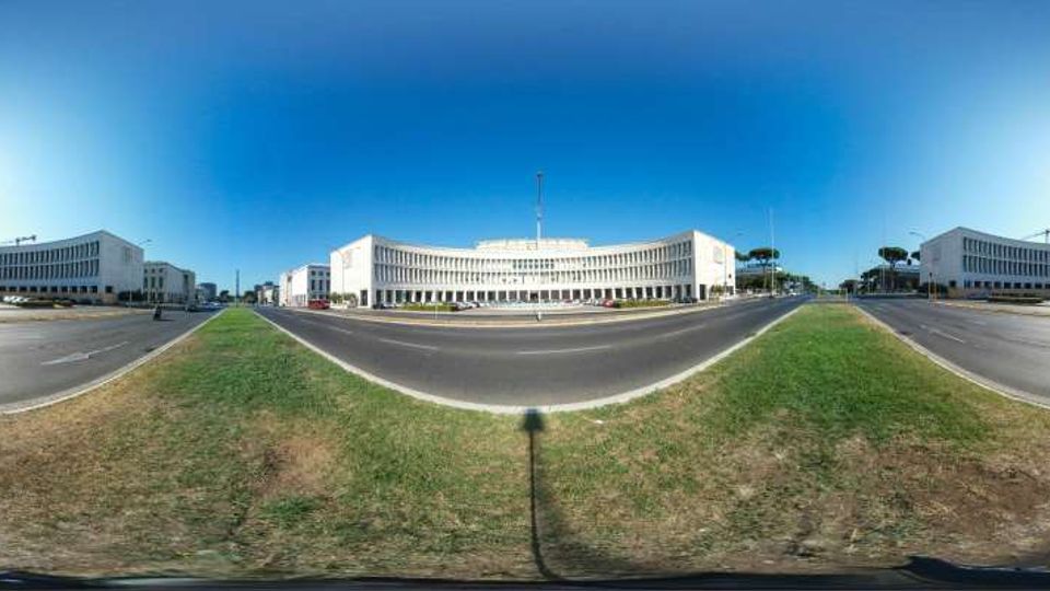 Vista panoramica a 360° di un ampio viale a più corsie con spartitraffico erboso e rotonda, circondato da grandi edifici moderni bianchi a forma curva con lunghe file di finestre. Sullo sfondo si vede un cielo sereno e alcuni alberi lungo il margine stradale.