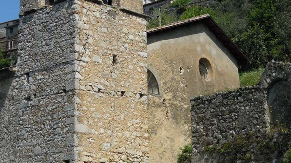 Campanile in pietra con aperture ad arco e campana visibile, accanto a una piccola chiesa con facciata intonacata e oculo. L’edificio sorge su un pendio erboso con muri in pietra e vegetazione, con case e alberi sullo sfondo sotto un cielo sereno.