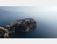 Veduta aerea di un borgo costiero fortificato con mura e bastioni in pietra, costruito su un promontorio roccioso a picco sul mare. Intorno si estende il mare aperto con costa e rilievi montuosi sfumati all’orizzonte.