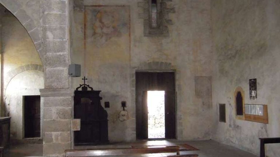 Interno di una piccola chiesa in pietra con soffitto in legno a capriate, panche in legno allineate e fonte battesimale in primo piano. Sul fondo si vedono una porta aperta, una stretta finestra e pareti intonacate con tracce di affreschi.