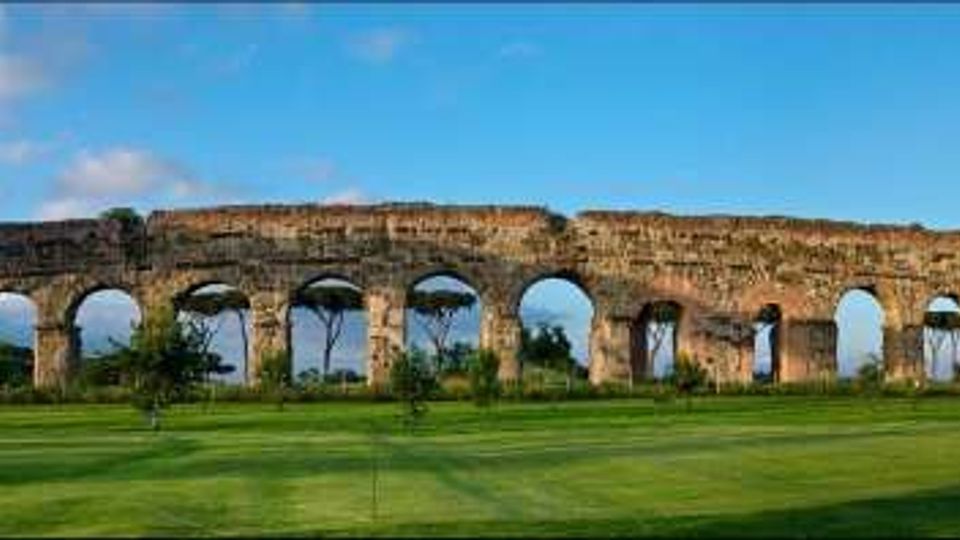 Panorama di un antico acquedotto romano in pietra con una lunga fila di arcate, esteso in orizzontale sopra un ampio prato verde. Sullo sfondo si vedono alberi ad alto fusto e un cielo azzurro con alcune nuvole.