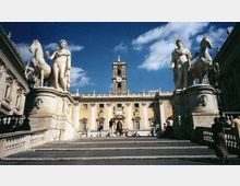 Scalinata monumentale che conduce a un palazzo storico con facciata simmetrica e torre dell’orologio centrale. Ai lati della scalinata ci sono grandi statue in pietra su piedistalli, con figure classiche e cavalli, sotto un cielo azzurro con nuvole.