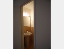 Piccolo bagno visto dalla porta, con lavabo a colonna e specchio illuminato da una lampada a parete. Le pareti sono rivestite a metà altezza con mosaico in piastrelle marroni e il resto è intonacato chiaro.