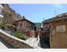 Vicolo in un borgo storico con case in pietra e tetti in coppi, percorso da una scalinata e da una rampa lastricata con muretti laterali. Sulla parete a destra è visibile un’insegna “Piazza della Fontana” e lampioni in ferro, con colline sullo sfondo.