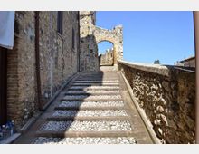 Scalinata in pietra con inserti di ciottoli bianchi che sale tra muri in pietra di edifici antichi, lungo un passaggio stretto. In cima si vede un arco in muratura collegato a una struttura di rovine, sotto un cielo limpido.