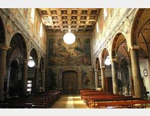 Interno di una chiesa con navata centrale fiancheggiata da arcate su colonne in pietra e file di panche in legno. Le pareti e la controfacciata sono riccamente affrescate, con soffitto a cassettoni decorato e grandi lampade sferiche sospese.