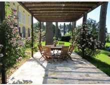 Pergolato in legno su una terrazza in pietra con tavolo rotondo e sedie da esterno, circondato da cespugli di rose. Sullo sfondo si vedono un prato con alti pini e il fianco di una villa gialla con finestre ad arco.