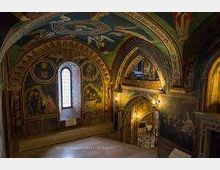 Interno di una chiesa o cappella storica con volta a botte e archi decorati da affreschi, una stretta finestra ad arco e un piccolo soppalco con scala. Pareti dipinte con motivi religiosi e illuminazione calda che mette in risalto le decorazioni.