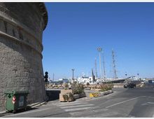 Vista di un’area portuale con strada asfaltata e banchina; a sinistra si vede la parete curva in pietra di una grande struttura circolare. Sullo sfondo sono ormeggiate navi e un grande veliero con alberi alti, con lampioni e fioriere lungo il bordo del porto.