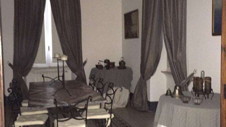Sala da pranzo in stile classico con pareti bianche, alte finestre con tende scure e cornici decorative sul soffitto. Al centro ci sono un tavolo con sedie in ferro battuto su un tappeto e, lungo le pareti, tavoli coperti da tovaglie con oggetti esposti.