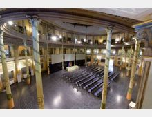 Ampia sala conferenze in un edificio storico con pianta ovale, colonne decorate e balconata perimetrale a due livelli. Al centro ci sono file di sedie rivolte verso un palco con schermi e tavolo, illuminate da luci a soffitto.