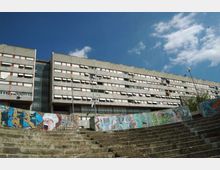 Grande edificio residenziale a più piani con facciata in pannelli grigi e lunghe file di finestre, con una torre vetrata centrale. In primo piano ci sono ampie gradinate in cemento e un muro basso coperto di graffiti, sotto un cielo azzurro con nuvole.
