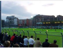 Stadio di calcio con campo in erba e recinzione metallica in primo piano, visto dalle gradinate con una tribuna coperta illuminata sul lato opposto. Sullo sfondo si vedono palazzi residenziali e un cielo nuvoloso al crepuscolo.