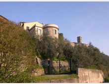 Edificio in pietra su un pendio collinare, con torretta cilindrica e finestre, circondato da ulivi e vegetazione. In primo piano si vedono un cancello metallico e muri di contenimento in pietra lungo una strada.