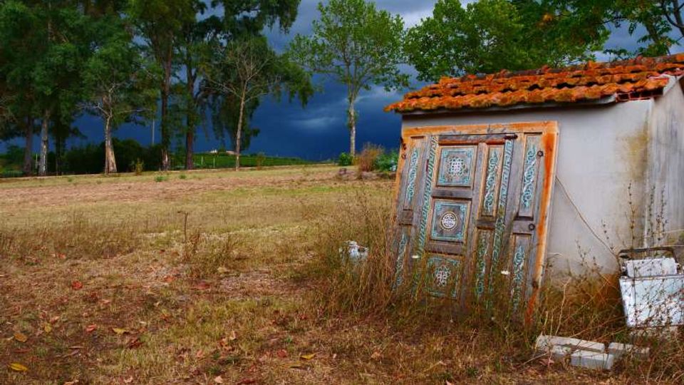 Piccolo edificio rurale intonacato con tetto in tegole rosse, con una porta decorata appoggiata all’esterno, ai margini di un campo erboso. Sullo sfondo si vedono alberi alti e un cielo scuro e nuvoloso.