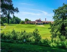 Ampio campo da golf con prati ondulati e un bunker di sabbia, circondato da alberi e vegetazione. Sullo sfondo si vede una club house in stile rustico con tetto in coppi, sotto un cielo azzurro sereno.