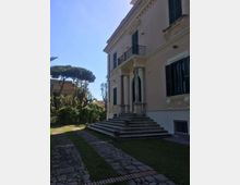 Facciata laterale di una villa storica color rosa con persiane scure, portico d’ingresso con colonne e una scalinata in pietra che conduce a una porta ad arco. Davanti si vede un vialetto lastricato tra strisce di prato e un giardino con alberi, con altri edifici sullo sfondo.