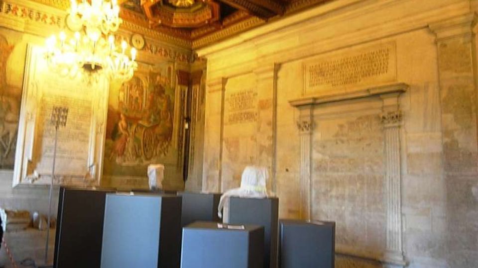 Interno di una sala storica con pareti in pietra incise, soffitto a cassettoni in legno dorato e grandi lampadari. Al centro sono presenti piedistalli scuri con frammenti scultorei e pavimento a motivi geometrici.