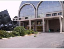 Ingresso di un edificio moderno con grande facciata vetrata ad archi e portico sorretto da pilastri, con cancelli metallici alla base. Davanti si vede un piazzale asfaltato con cespugli, una struttura nera con graffiti e un cestino verde.