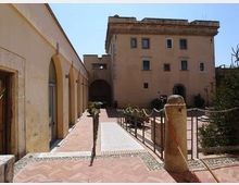 Cortile interno con pavimentazione in mattoni e percorso centrale, affiancato a sinistra da un porticato ad archi e di fronte da un edificio storico a più piani con finestre rettangolari. Sono presenti ringhiere e catene che delimitano le aiuole, con un grande pilastro in pietra in primo piano.