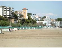 Spiaggia ampia con sabbia e una fila di cabine e recinzioni turchesi lungo il lungomare. Sullo sfondo si vedono edifici residenziali su una collina con palme e vegetazione, inclusa una torre in mattoni con tetto rosso.