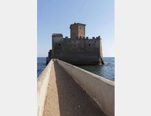 Fortezza in pietra sul mare con mura merlate e torre centrale, raggiungibile tramite una stretta passerella con parapetti in cemento. Il castello sorge su uno scoglio circondato dall’acqua, con cielo sereno sullo sfondo.