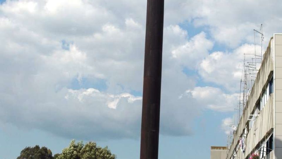 Terrazza pavimentata in cemento con parapetti bassi, dominata da un alto camino cilindrico scuro al centro. Sullo sfondo si vedono alberi e un edificio residenziale lineare con balconi e antenne sotto un cielo parzialmente nuvoloso.