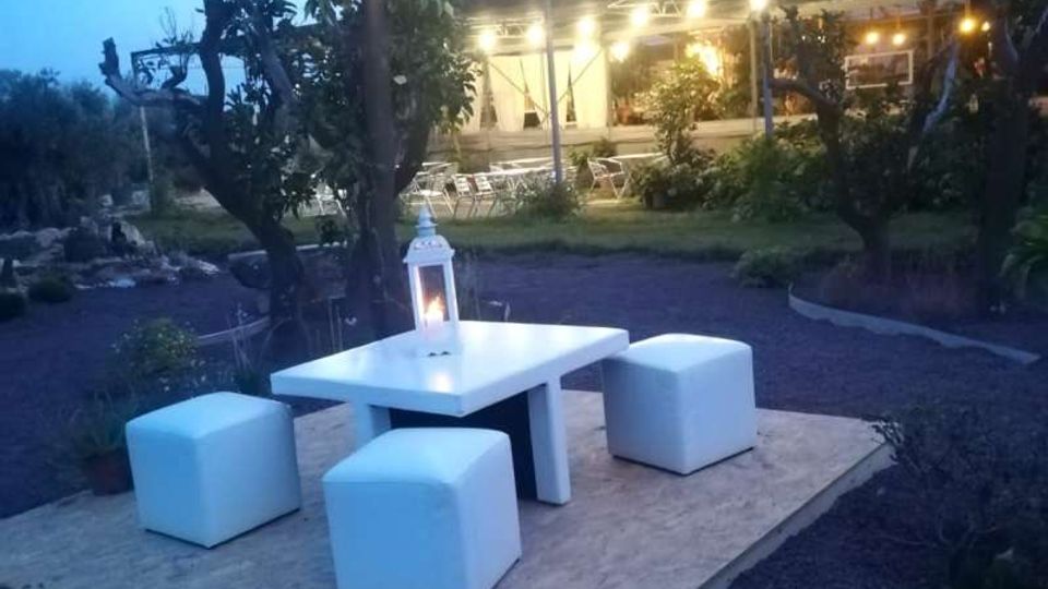 Area lounge all’aperto in giardino al crepuscolo, con tavolino bianco e quattro pouf su una pedana e una lanterna al centro. Sullo sfondo si vede un edificio con veranda illuminata e fili di lucine sospese tra gli alberi.