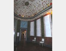 Interno di una sala circolare con pareti scandite da pannelli verticali e zoccolatura decorata, pavimento in legno e alcune sedie lungo la parete. Il soffitto a cupola è riccamente dipinto con motivi ornamentali e figure, con un’apertura che conduce a un corridoio di stanze.