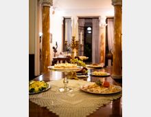 Sala da pranzo elegante con lungo tavolo di legno apparecchiato a buffet, piatti e alzate, e candelabro dorato al centro. L’ambiente è incorniciato da colonne in marmo e grandi finestre con tende chiare sullo sfondo.