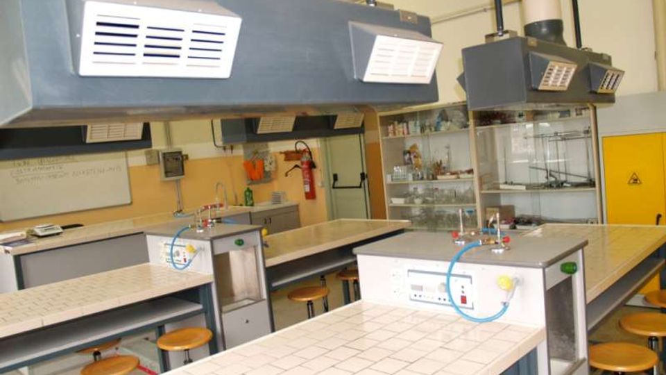 Laboratorio di chimica con banchi da lavoro rivestiti di piastrelle bianche, sgabelli e postazioni con rubinetti e attacchi per gas. Dal soffitto pendono grandi cappe aspiranti grigie, con armadi vetrati e scaffali sullo sfondo.