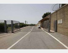 Strada asfaltata a due corsie con linea centrale tratteggiata, affiancata da recinzioni metalliche e da un lungo edificio industriale in mattoni con cancelli e cartelli. Lungo il lato destro è presente un piccolo veicolo parcheggiato e, sullo sfondo, si vedono vegetazione e pali della luce.