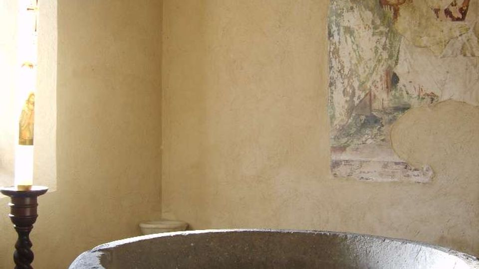 Interno di una piccola cappella con pareti in intonaco chiaro, un grande fonte battesimale in pietra al centro e un affresco religioso parzialmente rovinato sulla parete. A sinistra si vede una finestra con luce naturale e un portacandela alto vicino al pavimento in piastrelle.