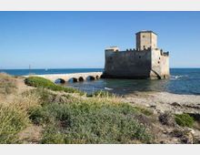 Fortezza in pietra sul mare con torre quadrata e mura merlate, collegata alla riva da un ponte in muratura con archi. In primo piano si vede una costa rocciosa con vegetazione bassa e, sullo sfondo, il mare aperto sotto un cielo sereno.