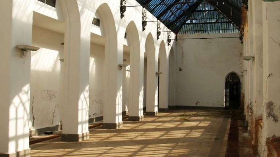Interno di un lungo corridoio con pareti bianche e una fila di grandi archi, coperto da un lucernario a falde in vetro e struttura metallica. La luce del sole filtra dal tetto creando ombre geometriche sul pavimento in cemento, con canaletta di drenaggio lungo un lato.