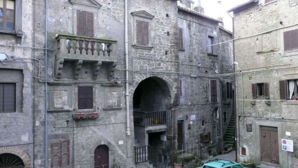 Facciate di edifici in pietra in un borgo storico, con finestre a persiane, balcone con balaustra e un grande arco passante al centro. Piccola piazzetta lastricata con scale laterali e un’auto parcheggiata vicino agli ingressi.
