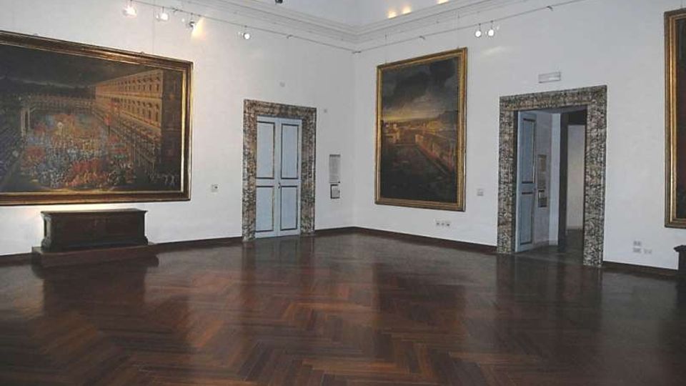 Sala museale con pareti bianche e pavimento in parquet scuro a spina di pesce, illuminata da faretti a soffitto. Grandi dipinti in cornici dorate sono appesi alle pareti e si vedono porte azzurre incorniciate in pietra che conducono ad altre stanze.