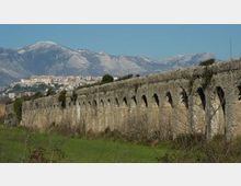 Lungo acquedotto in pietra con una serie di archi ripetuti, che attraversa un prato con vegetazione bassa. Sullo sfondo si vedono un centro abitato su una collina e alte montagne sotto un cielo sereno.