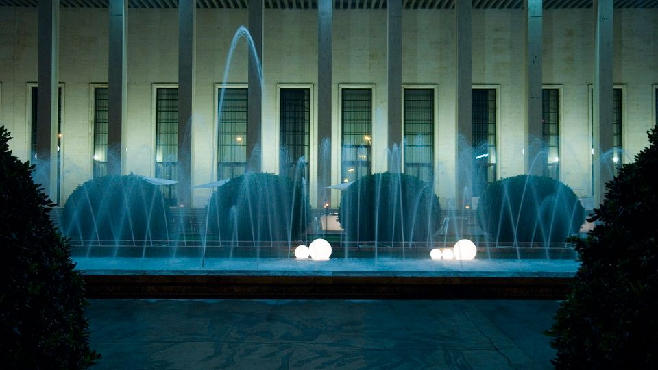 Fontana illuminata di notte con getti d’acqua davanti a un edificio monumentale con alte colonne e grandi finestre verticali. Nella vasca si vedono sfere luminose bianche e siepi potate, con un’ampia pavimentazione in primo piano.